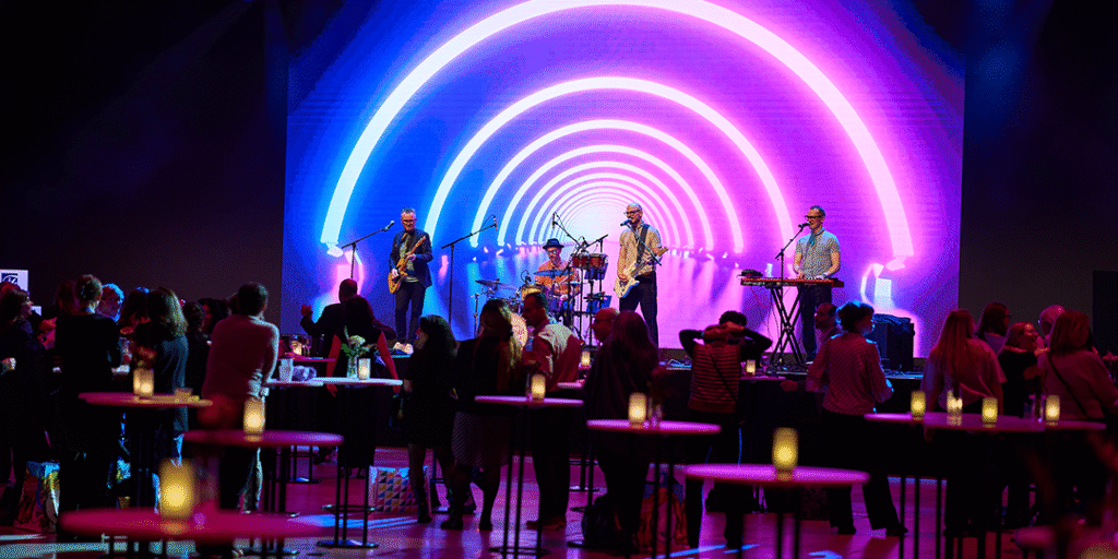 Liveband spelar inför publik, färgglad scenbakgrund i neon och ståbord med ljus under en kickoff i Victoriahallen på Stockholmsmässan