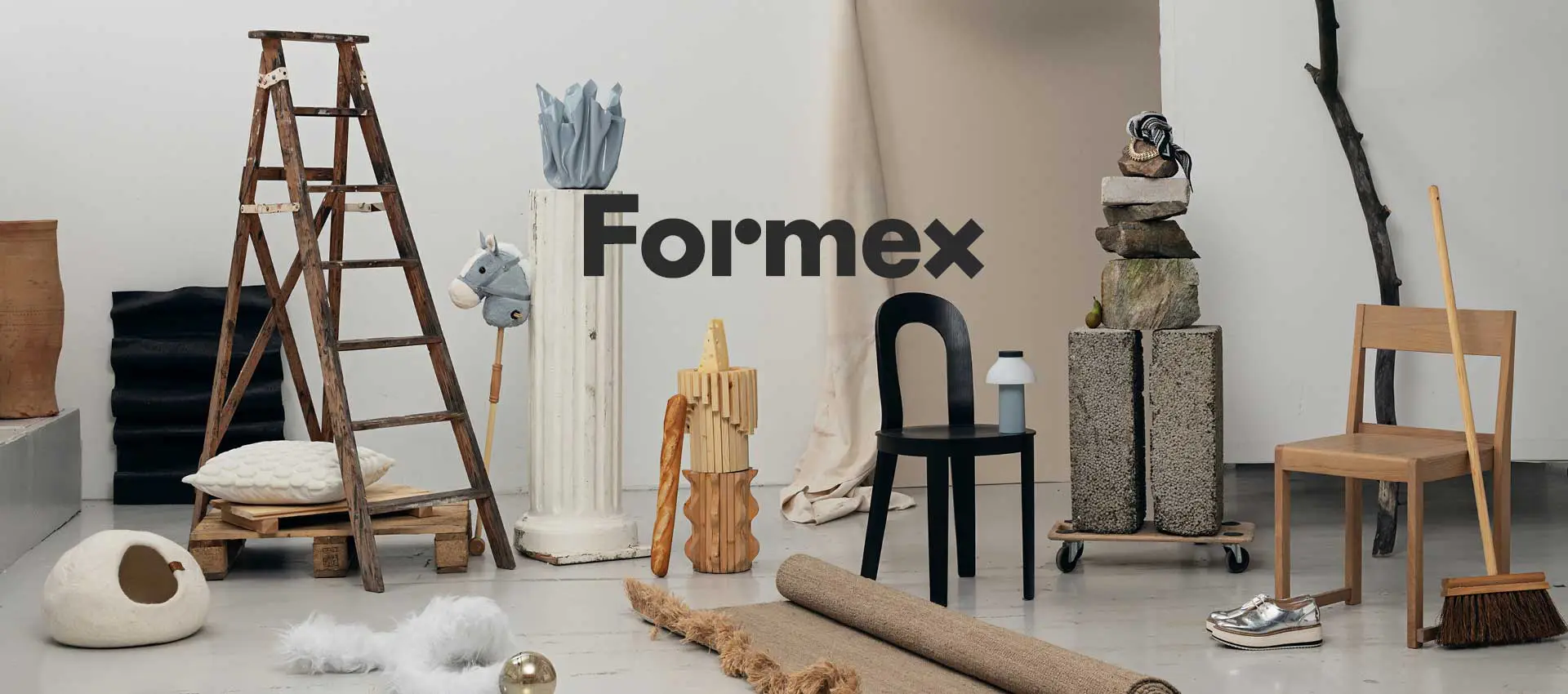 Formex 1 En stylad inomhusscen med olika inrednings- och designobjekt, där Formex‑logotypen syns i mitten; bland objekten finns en trästege, stolar, pedestaler, dekorativa föremål och naturmaterial arrangerade i ett kreativt stilleben.